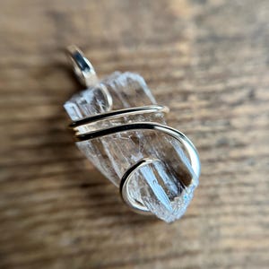Könnte beinhalten: Ein klarer Kristall-Anhänger, der mit goldfarbenem Draht umwickelt ist. Der Kristall hat eine lange, dünne Form mit einer spitzen Spitze. Der Anhänger hängt an einer goldenen Kette.