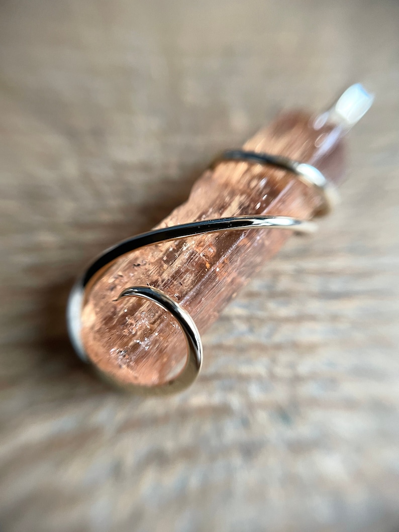 May include: A pink and brown crystal pendant wrapped in a gold wire. The pendant is on a wooden surface.