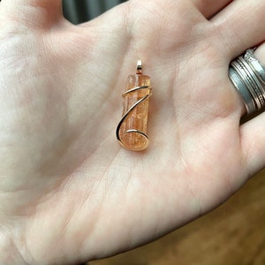 May include: A gold wire wrapped pendant featuring a natural orange-brown crystal. The crystal is long and thin, with a smooth, polished surface.