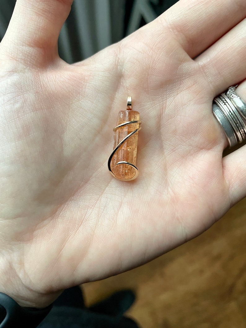 May include: A gold wire wrapped pendant featuring a natural orange-brown crystal. The crystal is long and thin, with a smooth, polished surface.