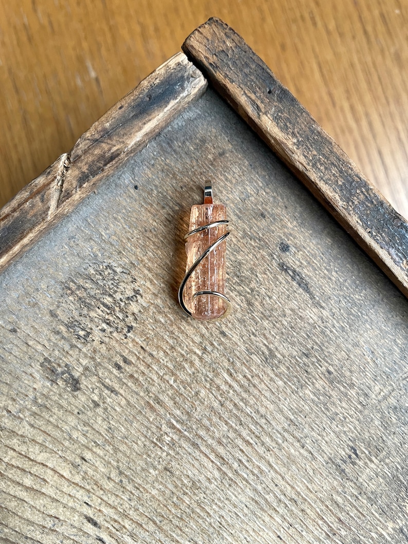 May include: A gold wire wrapped pendant featuring a natural, translucent, orange-brown crystal. The crystal is a long, thin, rectangular shape with a pointed end.