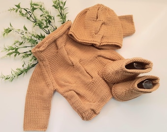 May include: A tan-colored knitted baby set, including a sweater, hat, and booties. The set is made of soft yarn and is displayed on a white surface. The sweater has long sleeves and a round neckline. The hat is a beanie style.