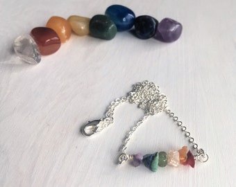 Può includere: Una collana con catena d'argento con un pendente a barra con una fila di piccole pietre preziose grezze colorate. Le pietre preziose sono disposte in uno spettro arcobaleno, con colori come rosso, arancione, giallo, verde, blu, viola e trasparente. Un insieme di pietre lucide degli stessi colori è sullo sfondo.