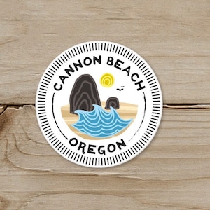 May include: A round sticker with a beach scene. The sticker features the text "Cannon Beach Oregon" in a circular design. The image includes a black rock formation, blue waves, a yellow sun, and a small bird.