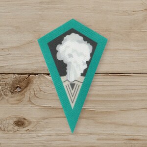 May include: A diamond-shaped patch with a teal border and a central design. The design features a white cloud-like shape above a gray and white geometric pattern. The background is a light wood grain.