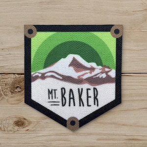 May include: A fabric patch featuring a mountain landscape with the text "MT. BAKER." The design includes a mountain range in shades of brown and white, set against a green and black background. The patch has a black border with brown circular accents.