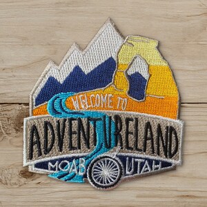 May include: Embroidered patch with the text "WELCOME TO ADVENTURELAND MOAB UTAH." The patch features a mountain range, a river, and a wheel design, with a color palette of blue, orange, yellow, and white.