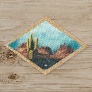 May include: Diamond-shaped patch featuring a desert landscape with a tall cactus and reddish-brown rock formations under a blue sky. The patch has a tan border and is set against a wooden background.
