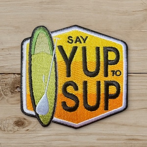 May include: Embroidered patch featuring a stand-up paddleboard and paddle. The patch is shaped like a hexagon with a yellow and black border. Text on the patch reads "SAY YUP TO SUP". The paddleboard is green and white.