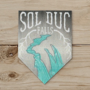 May include: A pennant-shaped patch with the words "SOL DUC FALLS" in white, stylized lettering. Below, a turquoise-colored illustration depicts a waterfall. The background is a gradient of gray and white, with a textured appearance.