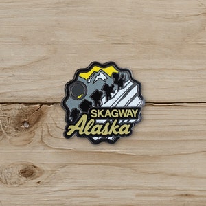May include: A black and yellow Skagway Alaska souvenir pin. The pin features a mountain range, a sun, and the words "Skagway Alaska" in a stylized font. The pin has a black border and is set against a wooden background.