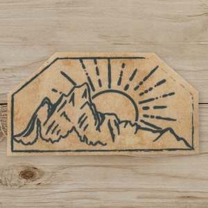 May include: A beige patch featuring a mountain range silhouette with a rising sun design. The mountains and sun rays are in a dark gray color. The patch has a unique, irregular shape, and is likely made of fabric.