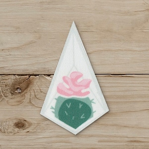 May include: A white, triangular fabric earring with a watercolor design of a pink flower and a green cactus. The earring is set against a wooden background.