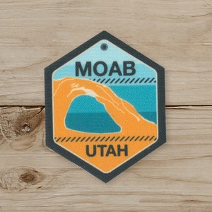 May include: A hexagonal patch featuring the text "MOAB" above an orange archway and the text "UTAH" below. The design includes blue and orange colors, with a black border and a small hole at the top for attachment. The patch is set against a wooden background.