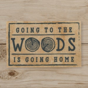 May include: A rectangular sign with the text "GOING TO THE WOODS IS GOING HOME" in dark blue lettering. The sign has a tan background with a textured appearance, and a dark blue border. The word "WOODS" features a spiral design.