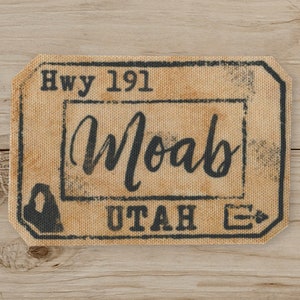 May include: A rectangular beige patch with the words "Hwy 191 Moab UTAH" in black, distressed lettering. The patch has a vintage, worn look, with a border and a small graphic of a mountain and an arrow.