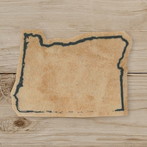 May include: A tan fabric patch in the shape of the state of Oregon, with a dark blue outline. The patch has a distressed, vintage appearance, with a slightly faded look. The background is a light wood grain.