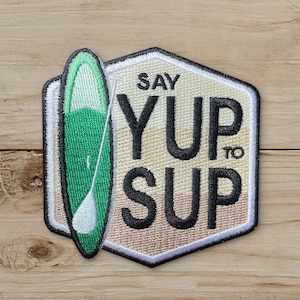 May include: Embroidered patch featuring a green and white paddleboard with a paddle. The patch is in a hexagon shape with the words "SAY YUP TO SUP" in black lettering. The patch has a white border and is on a wood grain background.