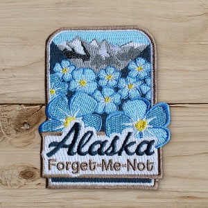 May include: Embroidered patch featuring the word "Alaska" and "Forget-Me-Not" in a rectangular shape. The patch has a light blue background with blue flowers and a mountain range. The patch is bordered with a brown trim.