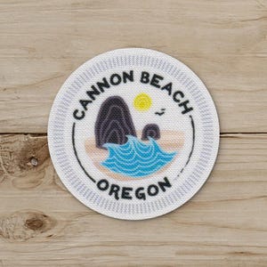 May include: A round patch featuring a coastal scene with the text "Cannon Beach Oregon". The design includes a dark rock formation, blue waves, a yellow sun, and a small bird. The patch has a white border with a textured pattern.