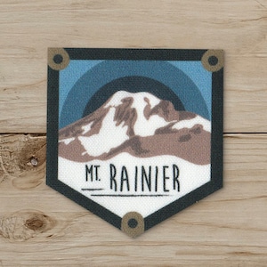 May include: A patch featuring a stylized illustration of Mount Rainier. The design includes a mountain with brown and white details, set against a blue and teal sky. The text "MT. RAINIER" is displayed in black, within a black-bordered, shield-shaped patch.