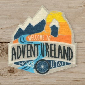 May include: A rectangular patch with the words "WELCOME TO ADVENTURELAND MOAB UTAH" in black and blue. The patch features a mountain range, a river, and a bicycle wheel design, with orange and yellow sunset colors.