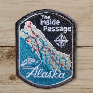 May include: Embroidered patch featuring a map of Alaska's Inside Passage. The patch has a black border and background with white text that reads "The Inside Passage" and "Alaska". The map is in shades of blue and gray, with red accents.