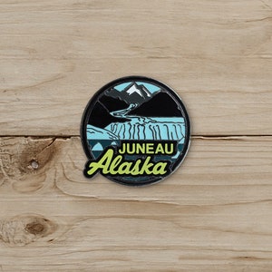 May include: A round enamel pin featuring a scenic view of Juneau, Alaska. The design includes a waterfall, mountains, and the text "Juneau Alaska" in a green and white font. The pin has a black border and a light blue background.