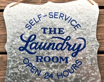 May include: A galvanized metal sign with a rope hanger. The sign has a decorative edge and features the words "SELF-SERVICE THE LAUNDRY ROOM OPEN 24 HOURS" in dark blue lettering. The sign is designed for a laundry room.