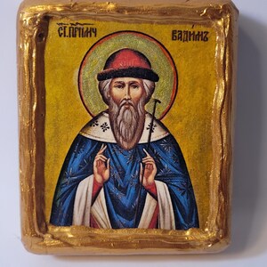 May include: A hand-painted icon of Saint Sergius of Radonezh, a Russian Orthodox saint. The icon is painted on a wooden panel and features a gold background with a halo around the saint's head. The saint is depicted with a long white beard and a blue robe. The icon is framed in a gold frame.