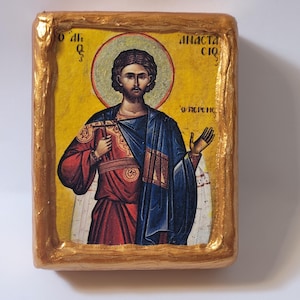 May include: A hand-painted icon of a saint with a gold background and a gold frame. The saint is wearing a red and blue robe and holding a cross. The icon is inscribed with the text "Ο άγιος" and "Διάστασις".