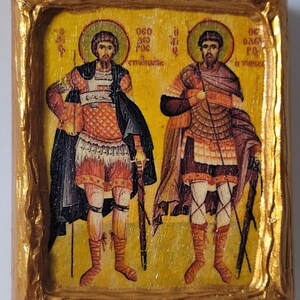 Pode incluir: Um ícone emoldurado a ouro que retrata duas figuras em trajes militares, cada uma segurando uma lança. As figuras estão em pé sobre um fundo amarelo com detalhes dourados. O texto "ΘΕΟ ΔΟΞΑ" está escrito acima de cada figura.