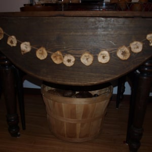 May include: A dark wood table with a garland of dried apple slices hanging from the edge. The table is set with a wooden basket filled with pine cones.