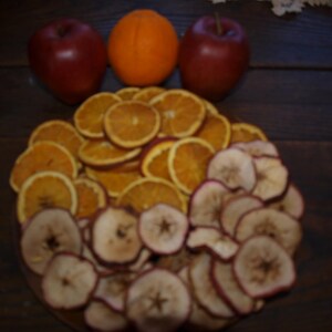 May include: A bowl of dried fruit slices, including orange and apple, with three whole apples in the background. Two of the apples are red and one is orange.