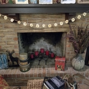 May include: A brick fireplace with a white paper flower garland hanging above the mantel. There are red candles on a black metal candle holder in front of the fireplace. There are three vintage tin boxes on the hearth, one with the text "Bless Our Home".