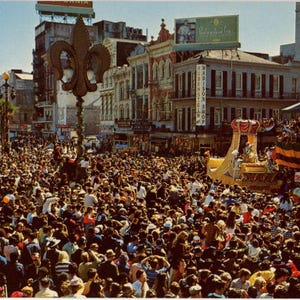 May include: Vintage postcard depicting a crowded street scene during Mardi Gras in New Orleans. A large crowd of people fills the street, with buildings and a parade float visible in the background. A fleur-de-lis is prominent.