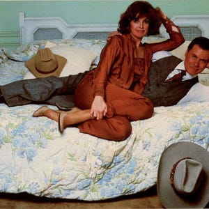 May include: A vintage photograph of a couple on a bed with a floral patterned quilt. The woman wears a rust-colored outfit, and the man is in a suit. A cowboy hat rests on the bed. Lamps and a nightstand are visible.
