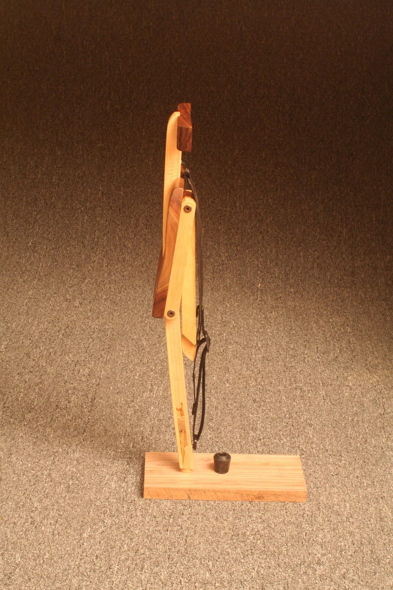 May include: A wooden folding chair with a black strap. The chair is folded and standing on a wooden base.