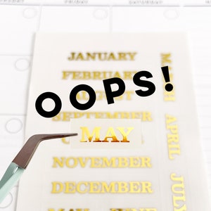 May include: A sheet of gold foil stickers with the months of the year printed on them. The word "OOPS!" is written in black letters on top of the sheet. A pair of tweezers is holding a "MAY" sticker.