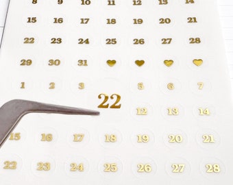 May include: Gold calendar stickers with numbers 1 through 31, hearts, and the number 22. The stickers are on a white sheet of paper.