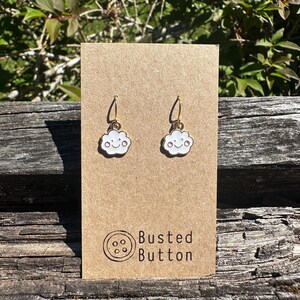 May include: A pair of gold-toned hook earrings featuring white enamel cloud charms with a smiling face and pink cheeks. The earrings are attached to a rectangular brown card printed with the text Busted Button and a circular button icon.