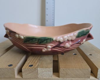 May include: A ceramic, oval-shaped planter with a peach-colored interior and a reddish-brown exterior. The planter features decorative floral and leaf accents in green and pink. The planter is resting on a wooden pallet.