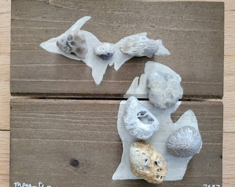 May include: A white painted outline of the state of Michigan on a brown wooden board. The outline is decorated with various small, light brown and gray stones. The board is signed "Megan Shaw" and dated "2022".