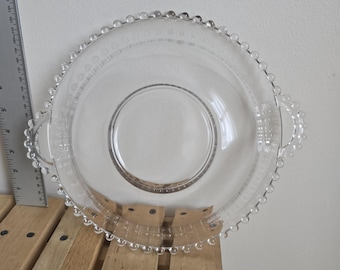 May include: A clear glass serving dish with a circular design. The dish has a beaded edge and two handles. The center of the dish is a smaller circle. The dish is sitting on a wooden surface.