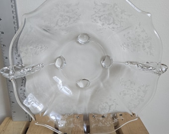 May include: Clear glass serving dish with a scalloped edge and etched floral design. The dish has a circular center with four small, raised feet and two handles. A ruler shows the dish is approximately 10 inches in diameter.