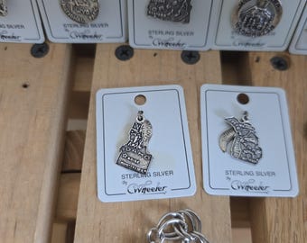 May include: Sterling silver charms featuring state shapes, including Idaho and Montana, displayed on white cards. The charms are attached to a silver chain. The cards are labeled "Sterling Silver" and signed "CWheeler".