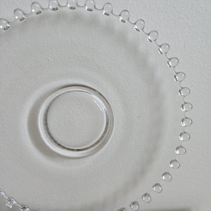 May include: Clear glass plate with a beaded edge and a circular center. The plate is transparent, allowing the white surface beneath to show through. The beaded edge adds a decorative touch to the simple design.