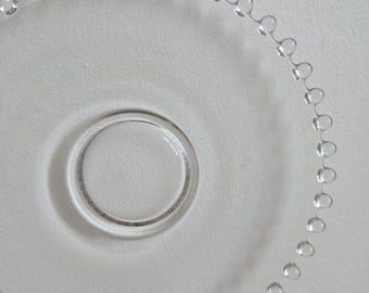May include: Clear glass plate with a beaded edge and a circular center. The plate is transparent, allowing the white surface beneath to show through. The beaded edge adds a decorative touch to the simple design.