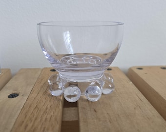 May include: A clear glass bowl with a rounded shape, resting on a base of clear, spherical accents. The bowl sits on a wooden surface. The bowl is designed to hold liquids or small items.