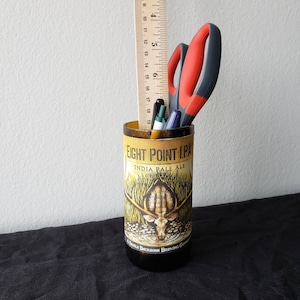 May include: A glass bottle with a label that reads "Eight Point IPA India Pale Ale" and features a drawing of a deer. The bottle is filled with pens, pencils, and a pair of scissors.